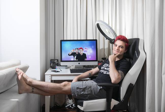 Foto mostra Felipe Neto, de cabelos vermelhos, sentado numa cadeira gamer, com a mão no queixo e um monitor e um ring light atrás dele