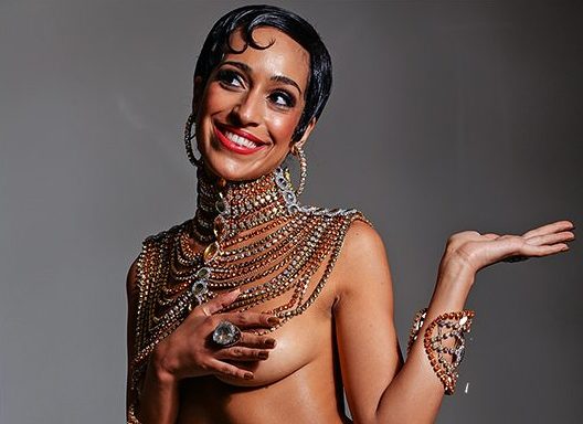 A atriz Aline Luna caracterizada como Josephine Baker, com cabelo curto e com gel, com mecha curva na testa, brincos de argola, muitos colares no pescoço e peito no coberto com a mão direita da atriz, que está sorrindo