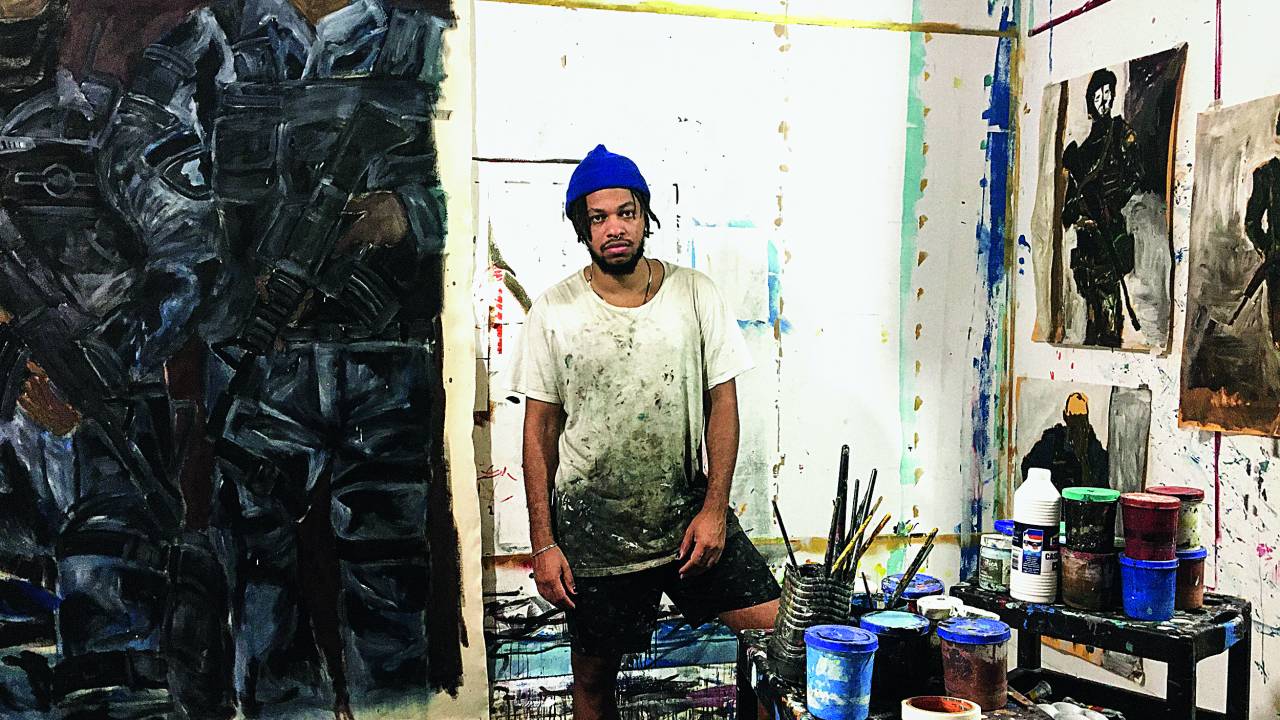 artista maxwell alexandre posa em seu atelier, há pincéis e telas espalhados pelo espaço