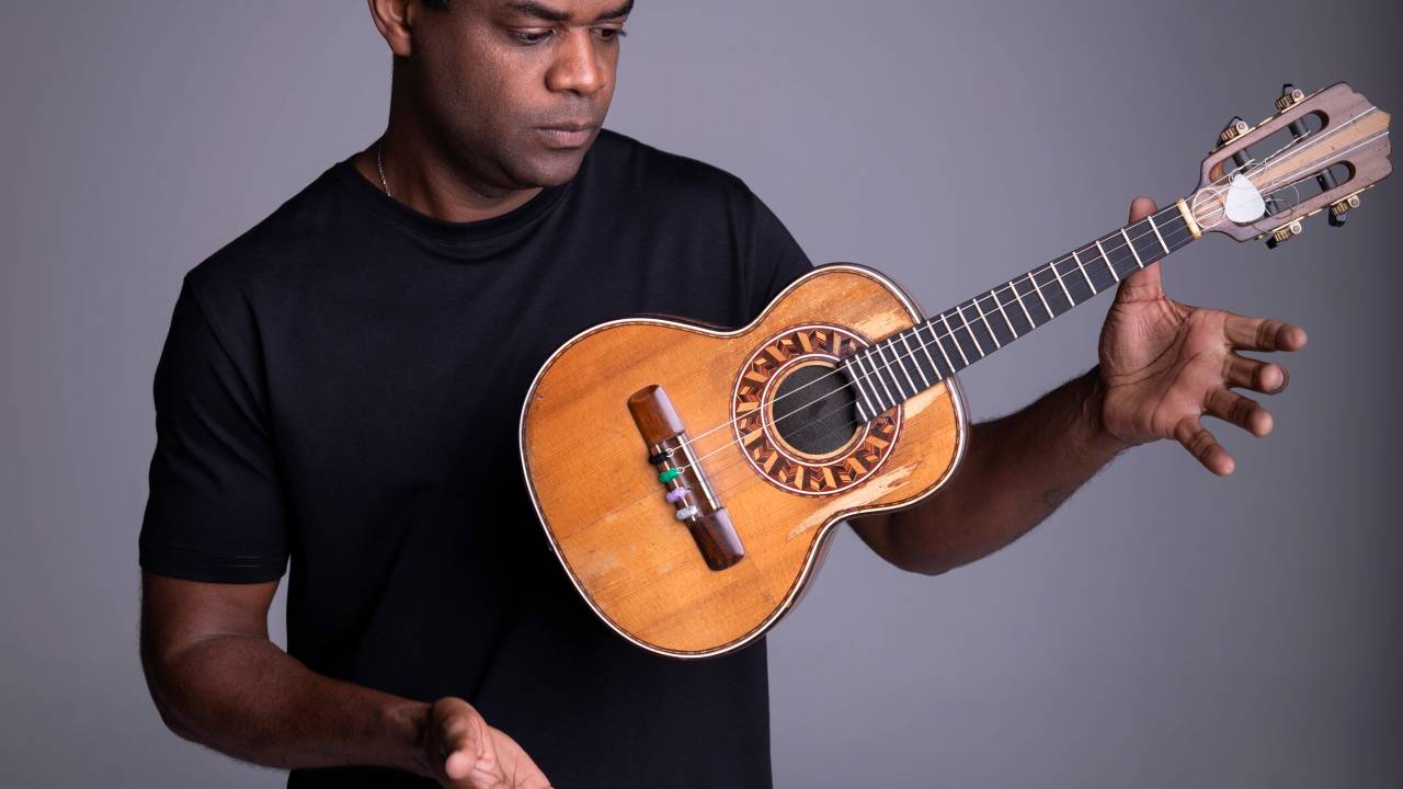 Pretinho da Serrinha de blusa preta jogando o cavaquinho para a frente, com as mãos em torno dele, mas sem encostar.