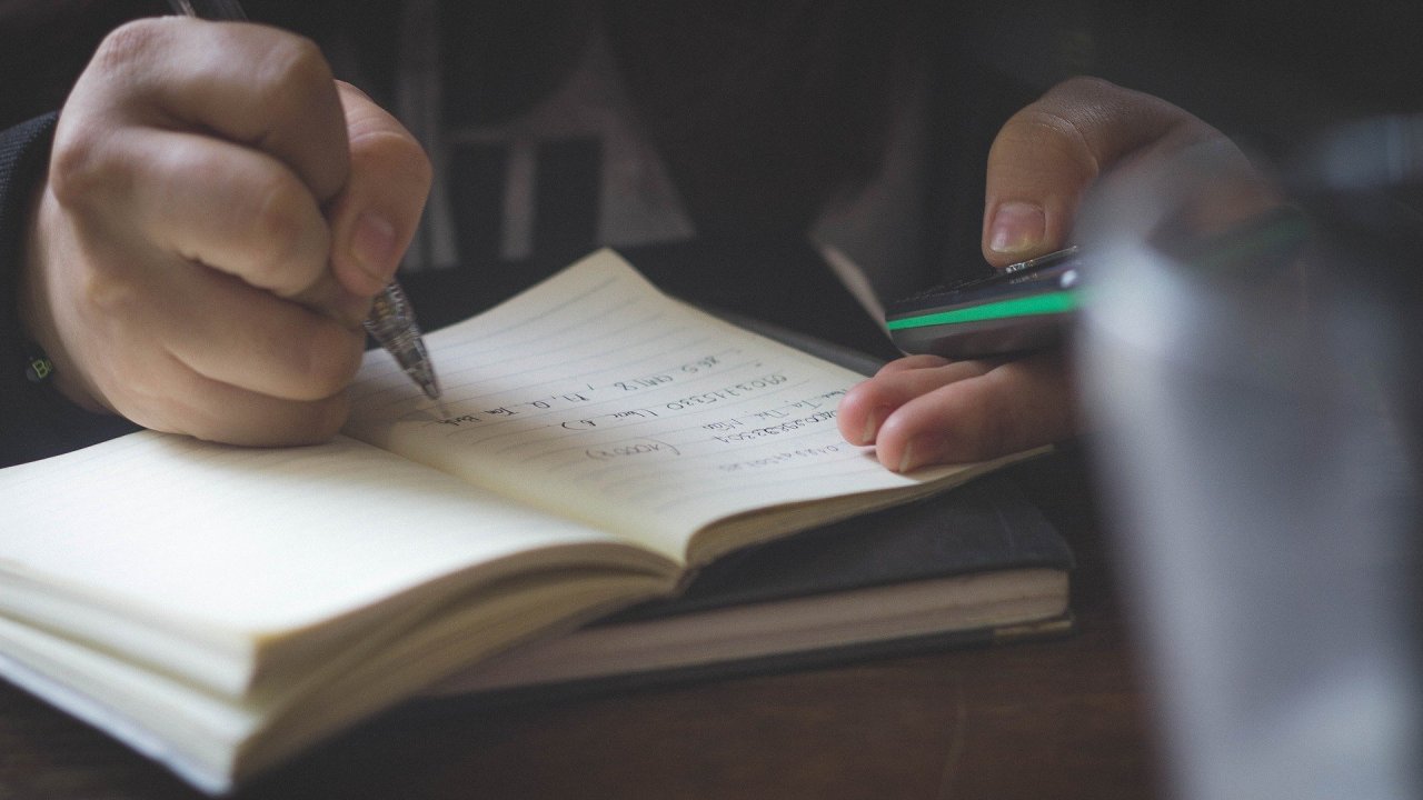 A imagem mostra uma pessoa escrevendo num caderno com um celular na outra mão