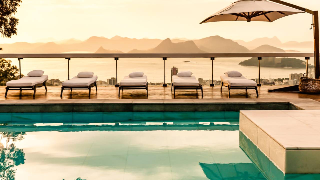 A imagem mostra a ampla piscina do Vila Santa Teresa, encarapitado no bucólico bairro de Santa Teresa, com espreguiçadeiras de estofado branco no entorno e vista privilegiada para prédios e para o mar carioca