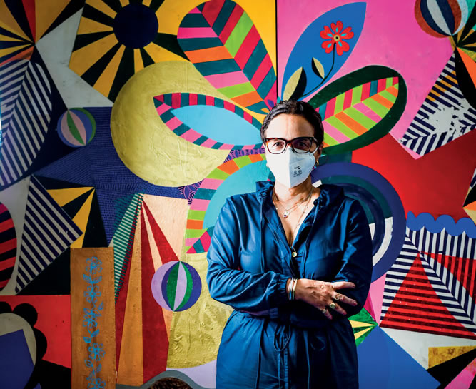 Beatriz Milhazes, de máscara N95, posa em frente a uma pintura multicolorida