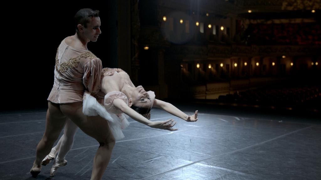 Num teatro vazio, bailarino segura bailarina num movimento de dança