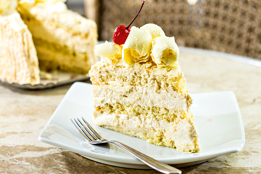 A imagem mostra uma fatia de torta de leite em pó, com cobertura de mussebranca, raspas de chocolate branco e cerejas