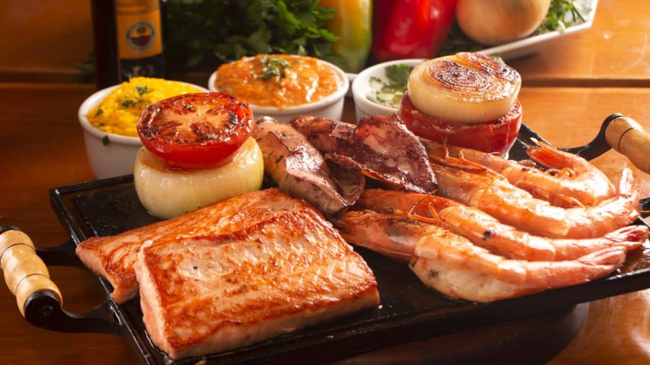 A imagem mostra uma chapa repleta de frutos do mar, com pescada amarela, lula e outros