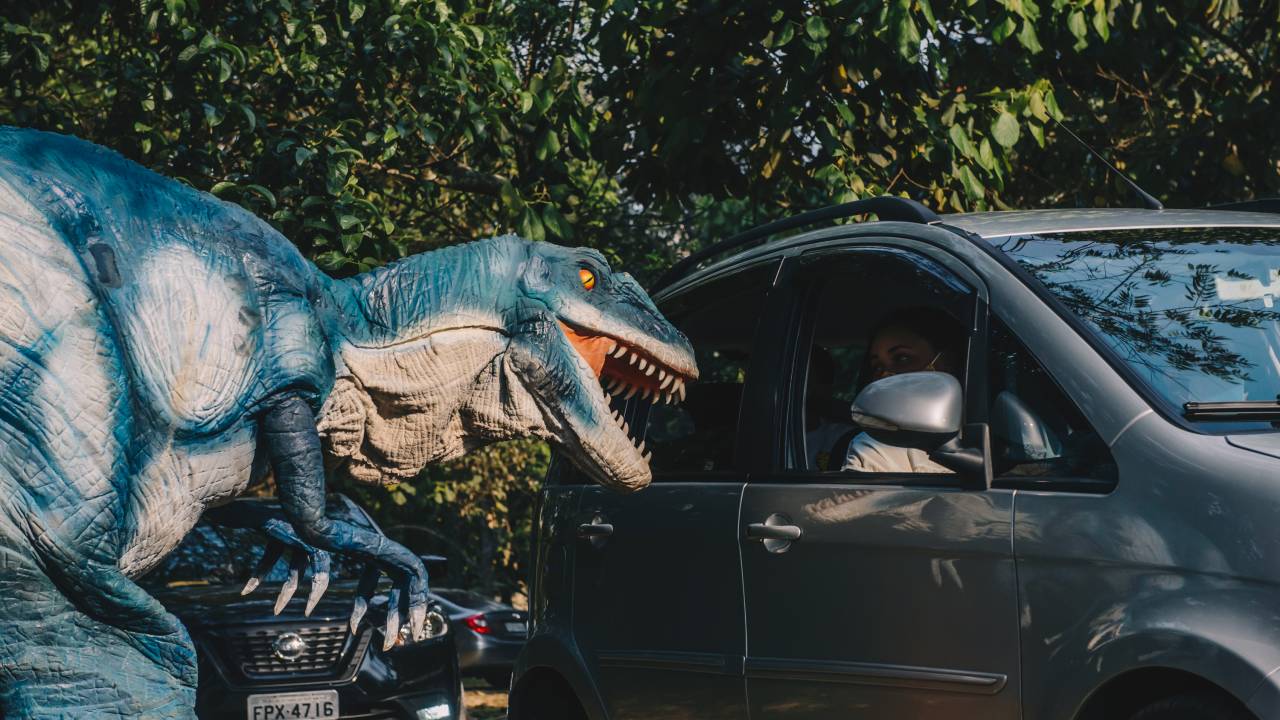 Réplica de tiranossauro rex ao lado de um carro