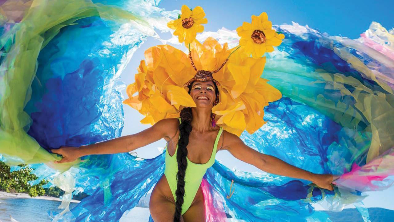 A imagem mostra a pernalta Raquel Potí de maiô verde-limão, vista de baixo para cima, sorrindo, com o céu azul atrás