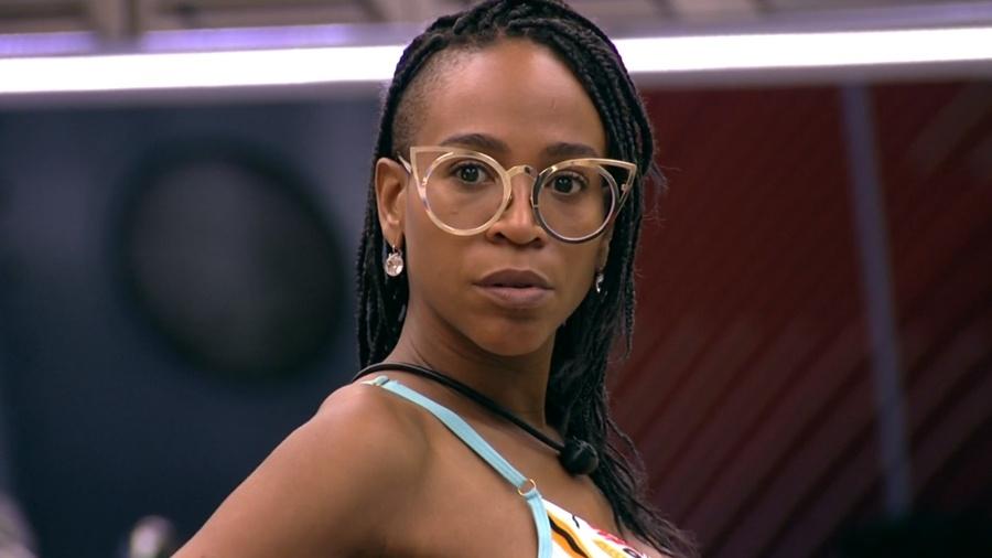 Mulher negra com tranças na altura dos ombros usando óculos modelo gatinho encara a câmera com expressão séria