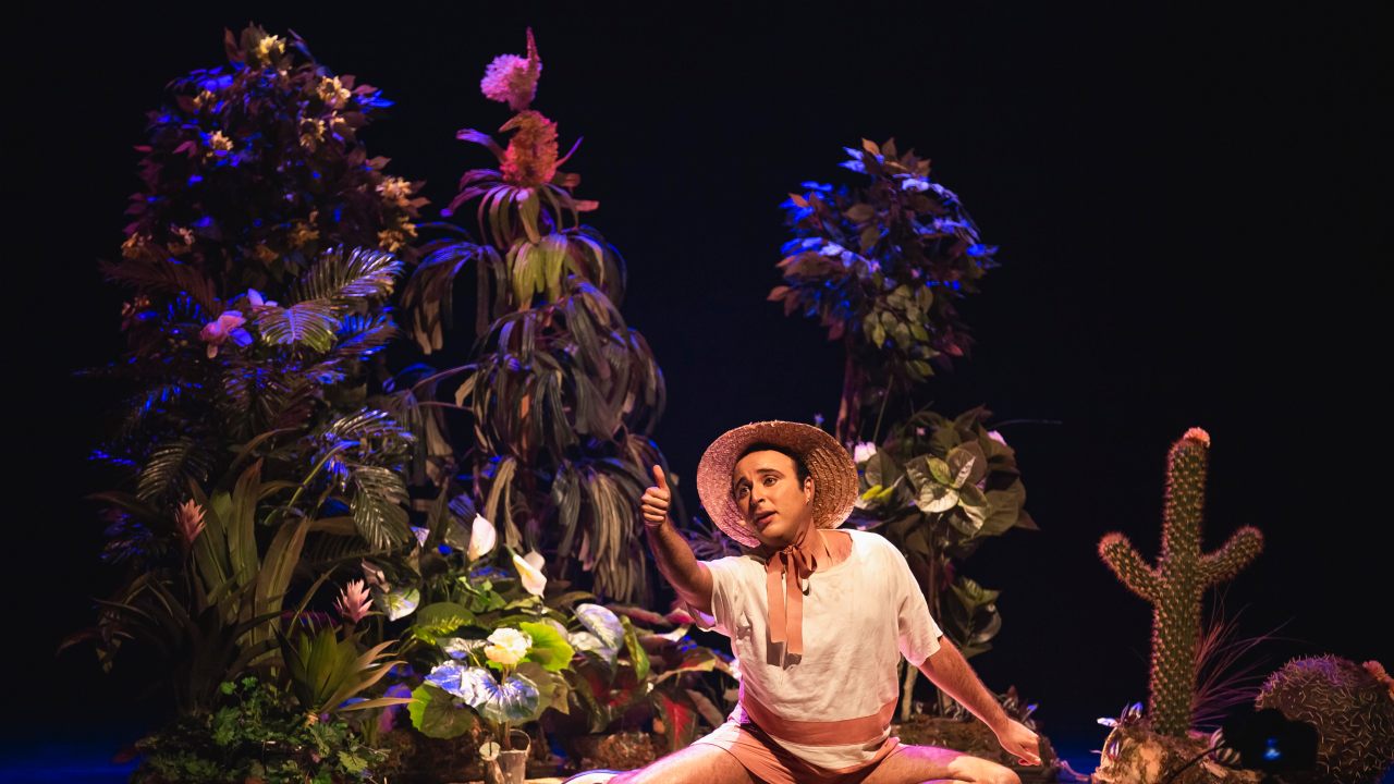 Ator Tauã Delmiro no palco com muitas plantas ao fundo