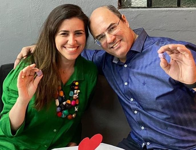 A imagem mostra o casal Helena e Wilson Witzel, governador afastado do Rio, sentado à mesa e sorrindo para a câmera