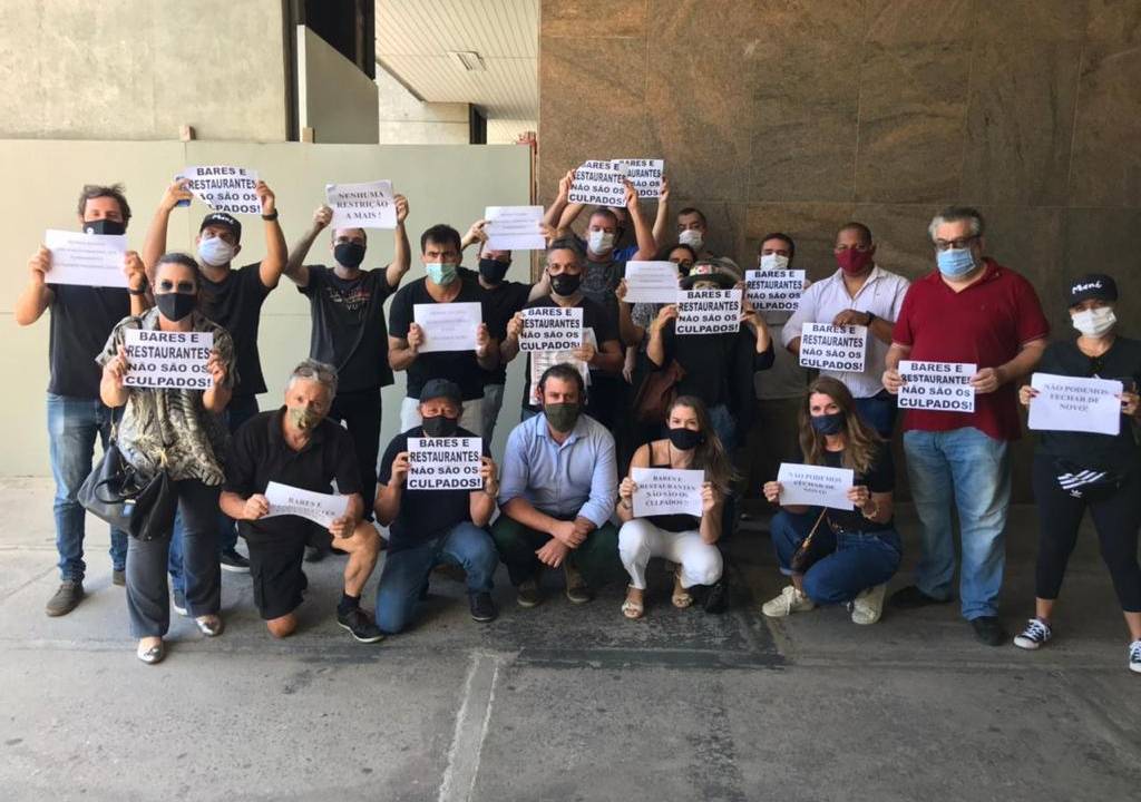 A imagem mostra um grupo de donos de restaurantes segurando placas de rpotesto contra as novas medidas restritivas na cidade, que os obrigam a funcionar com limitação de horario
