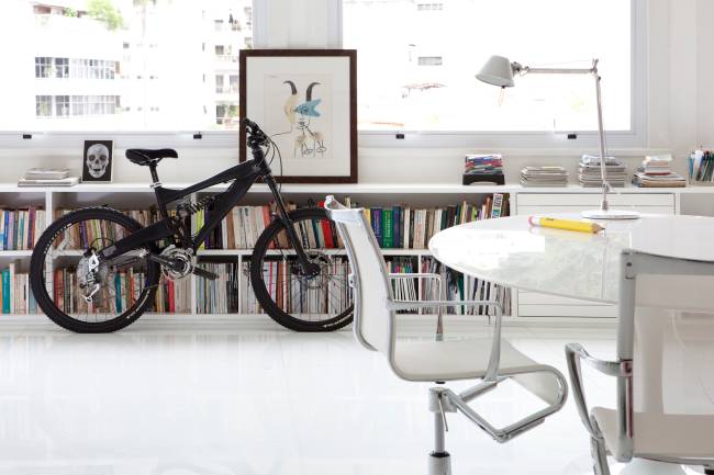 Bicicleta reinando na sala A imagem mostra uma bicicleta apoiada no aparador da sala. No primeiro plano uma mesa com uma cadeira.
