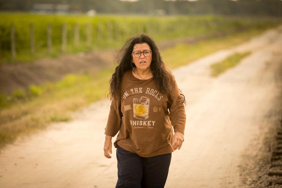 A imagem mostra a atriz Regina Casé como a personagem Lourdes, em Amor de Mãe, caminhando por uma estrada de terra