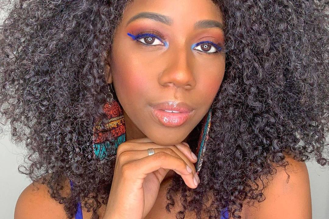 A imagem mostra a influencer Camilla de Lucas, olhando para câmera, de vestido azul e mão no queixo