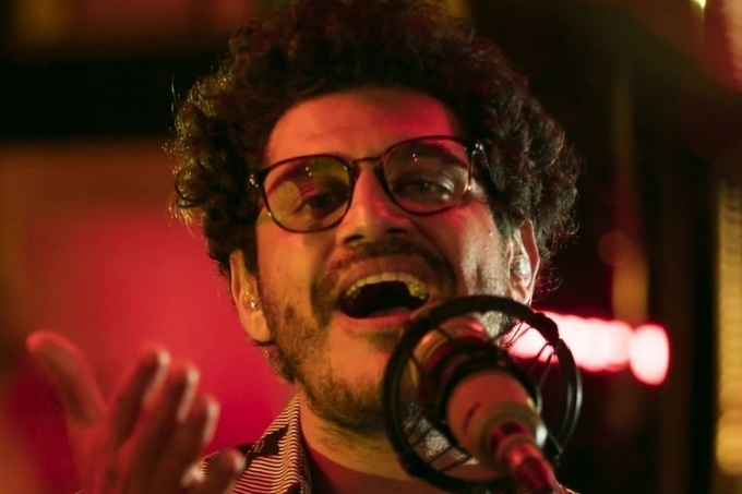 Criolo no palco, de óculos, cantando, com uma das mãos para cima