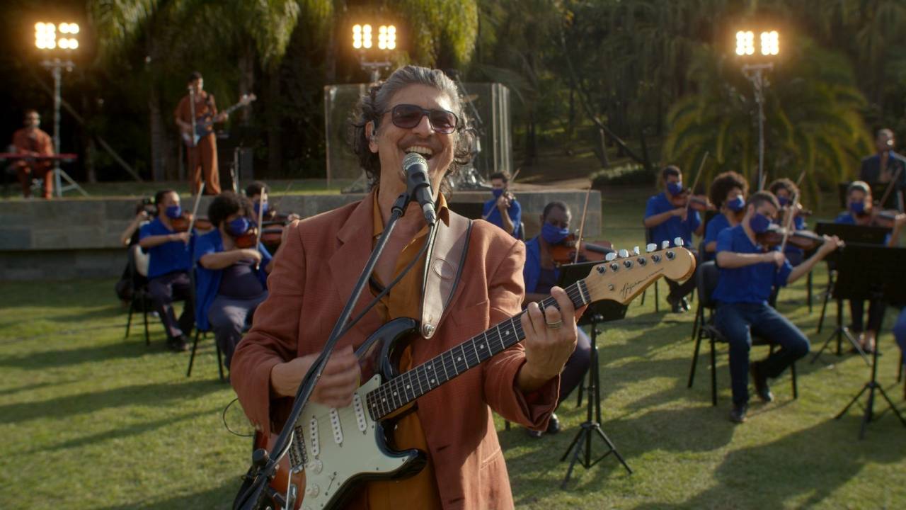 Pedro Luís ao ar livre, cantando e tocando guitarra, com os músicos da orquestra ao fundo