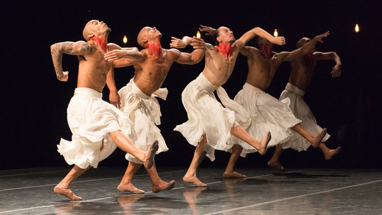 Cinco bailarinos no palco, usando espécie de saia, com os braços na frente do corpo