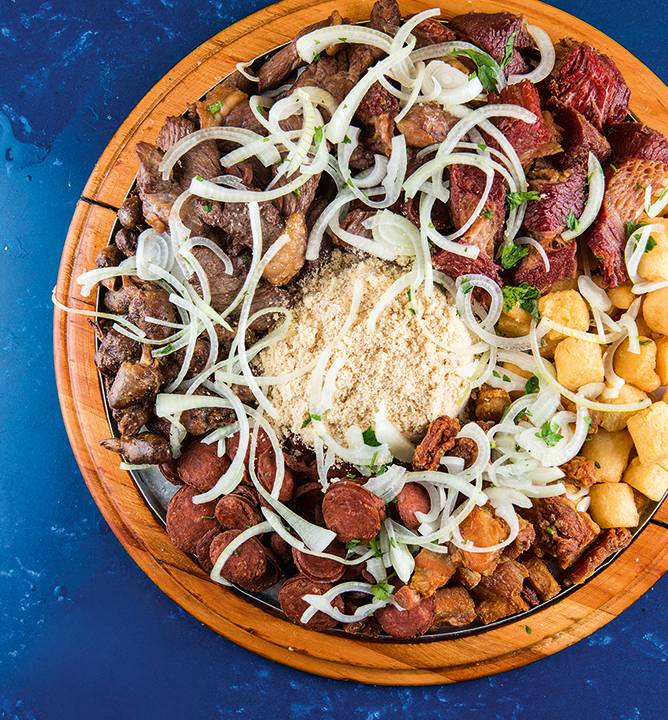 Foto mostra tábua com carnes, batata frita, farofa e cebola