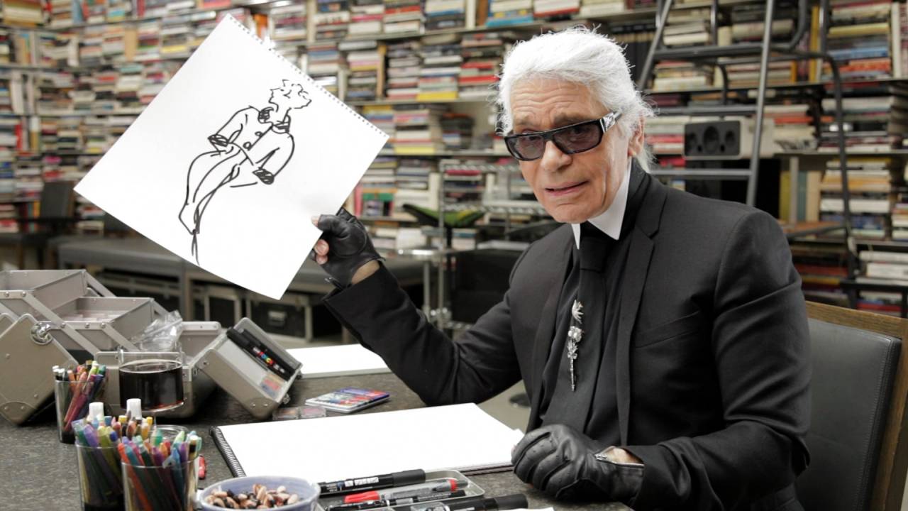 Foto do estilista alemão Karl Lagerfeld, morto em 2019, segurando um croqui. Ele está vestido com roupas pretas, inclusive luvas, e óculos escuros.