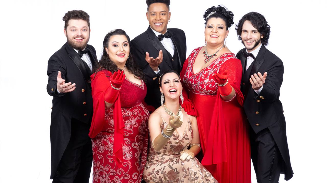 Os três solistas homens do espetáculo Esperança de Natal estão de fraque, duas mulheres estão de pé com vestidos vermelhos e uma mulher está sentada no centro da foto com vestido dourado