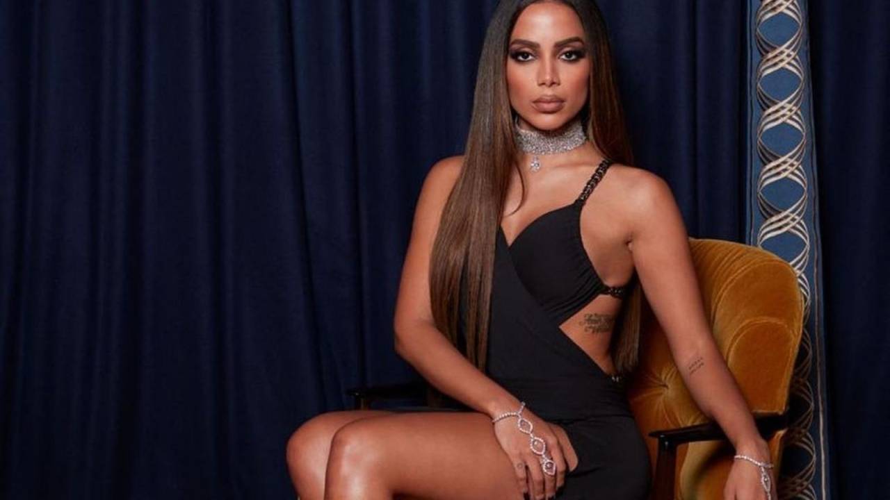 Foto mostra cantora Anitta sentada usando vestido preto, com as pernas à mostra