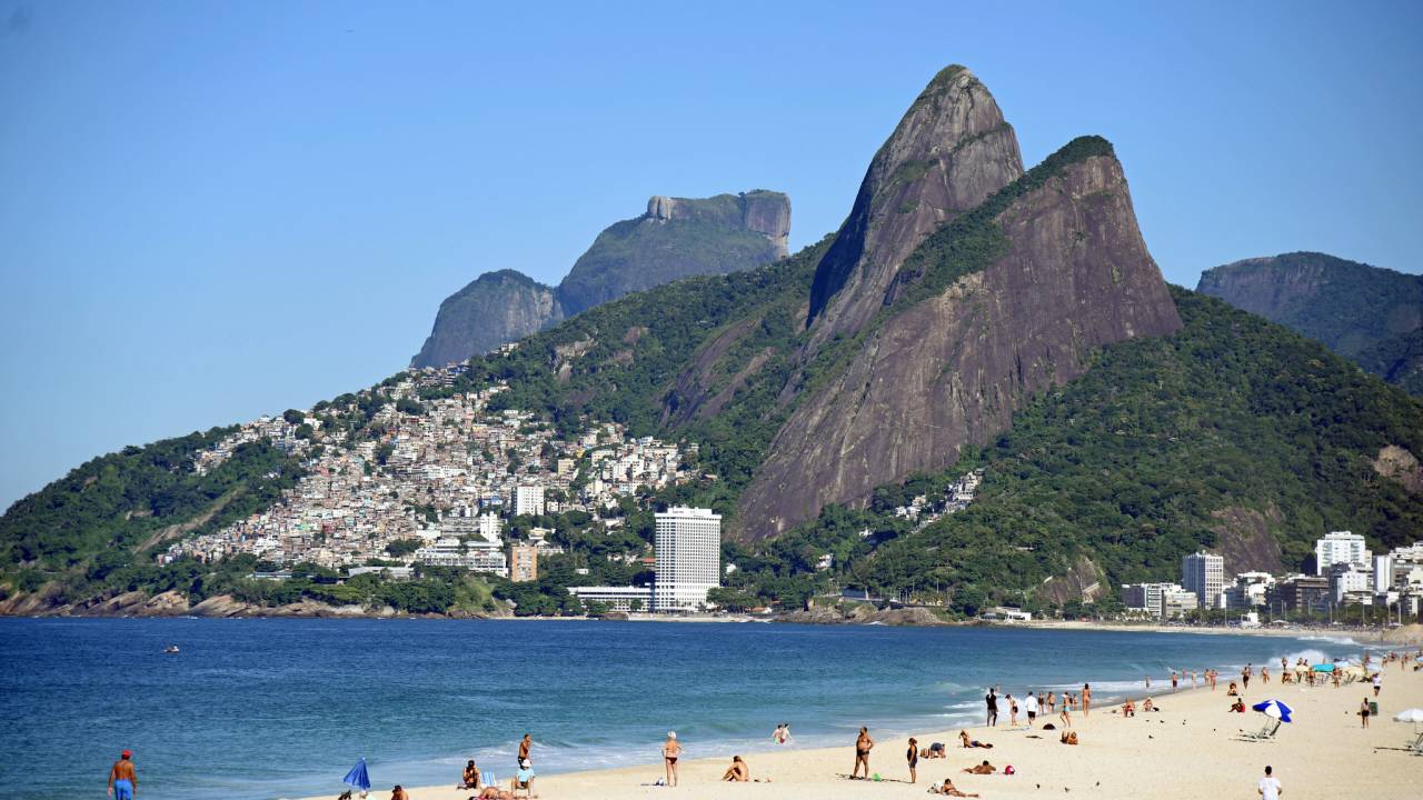 Um trecho da Praia de Ipanema, com o mar e a faixa de areia à direita, algumas pessoas sobre a areia. Ao fundo, o Morro Dois Irmãos.