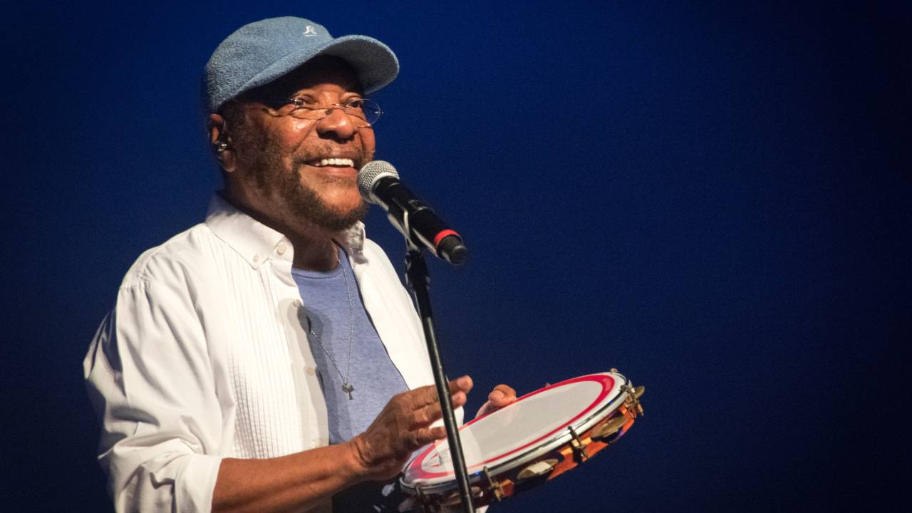 Martinho da Vila toca pandeiro no palco. Ele está de frente para o microfone. e veste blusa azul. camisa de manga comprida branca e boné azul.