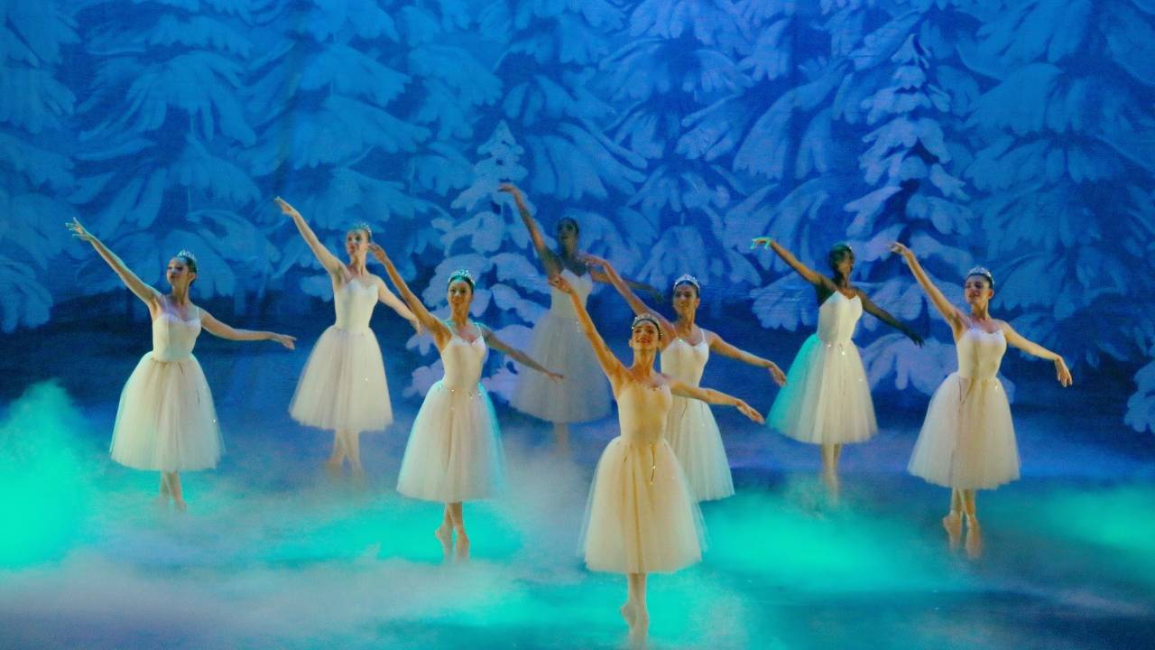 Bailarinas com as perna direita levantada, usando roupa branca com saia. Ao fundo, um cenário com pinturas de pinheiros brancos e luz azul.