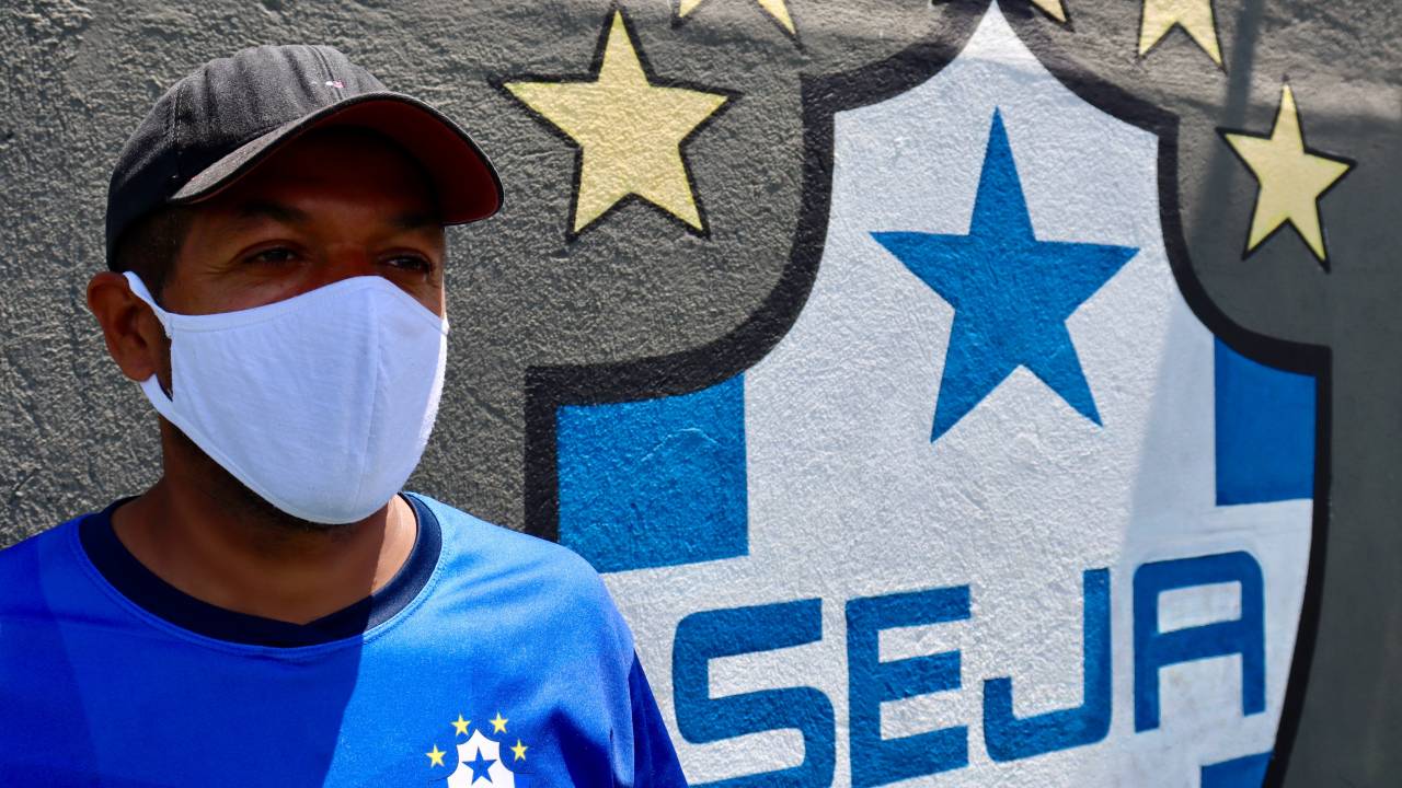 Foto mostra homem de camisa azul das elação brasileira, máscara facial branca e boné em frente a um brasão azul e branco,com uma estrela azul no topo e estrelas amarelas ao redor escrito SEJA