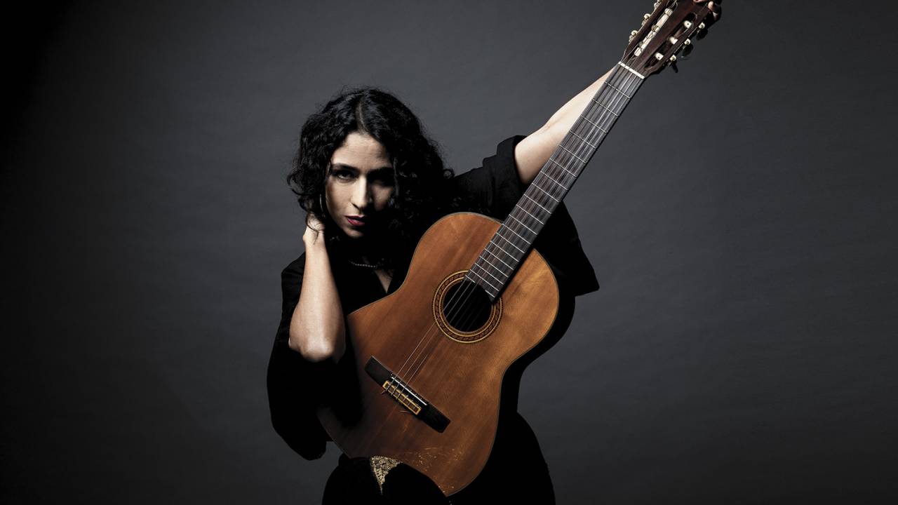 Marisa Monte com vestido preto segura violão e aponta o braço do instrumento para seu lado direito. Ela mira a câmera com olhar desafiador.