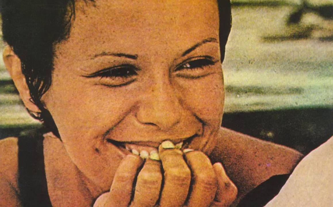 Elis Regina está sorrindo e tem a mão apoiada na boca cobrindo parte do sorriso. Ela usa cabelo curto e está olhando para o lado direito da foto.
