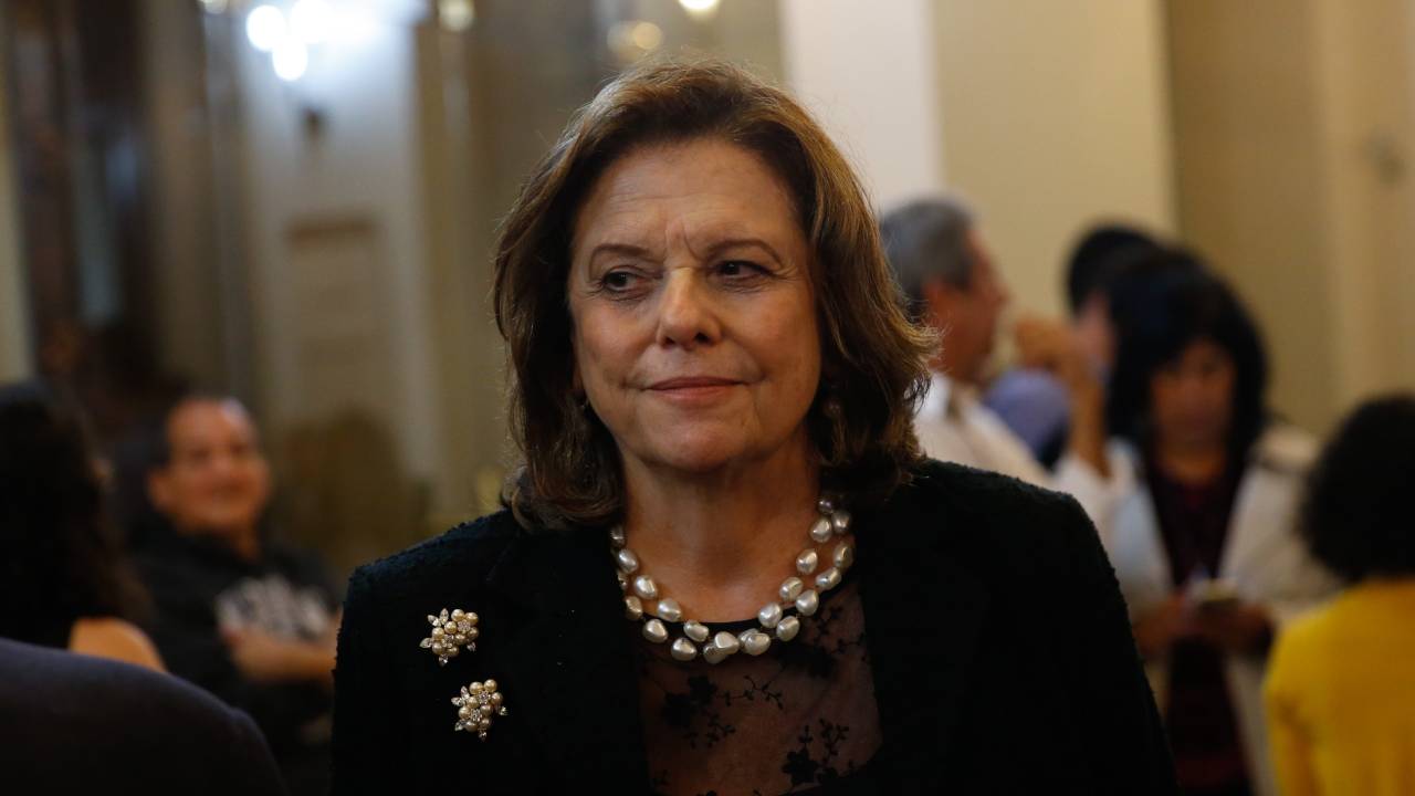 A gestora cultural Helena Severo está de paletó preto, blusa preta com detalhe de renda e colar nacarado de duas voltas. Ela tem os cabelos castanho-claros lisos na altura dos ombros e está olhando para a esquerda da foto.
