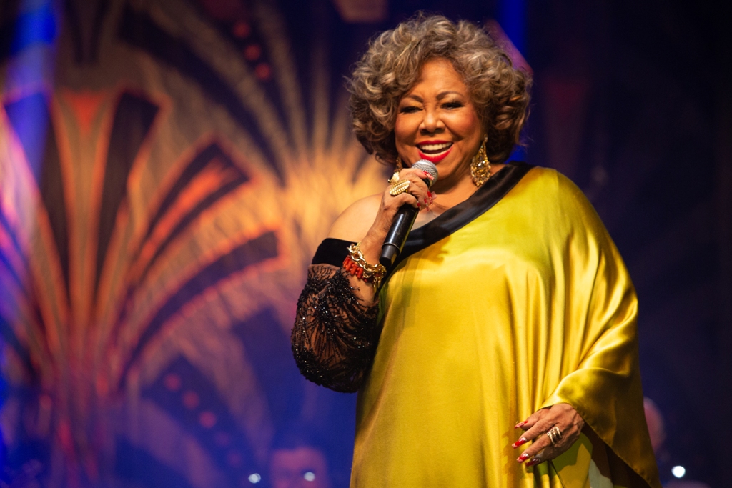 Alcione está cantando ao microfone, com vestido de cetim amarelo e uma das mangas preta e bordada