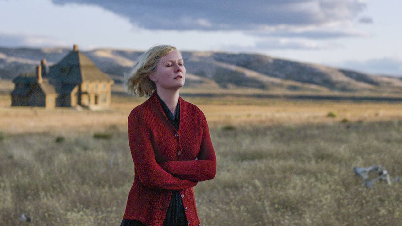A atriz Kirsten Dunst em um campo gramado. Ela usava casaco vermelho, saia preta, está com os braços cruzados e os cabelos ao vento