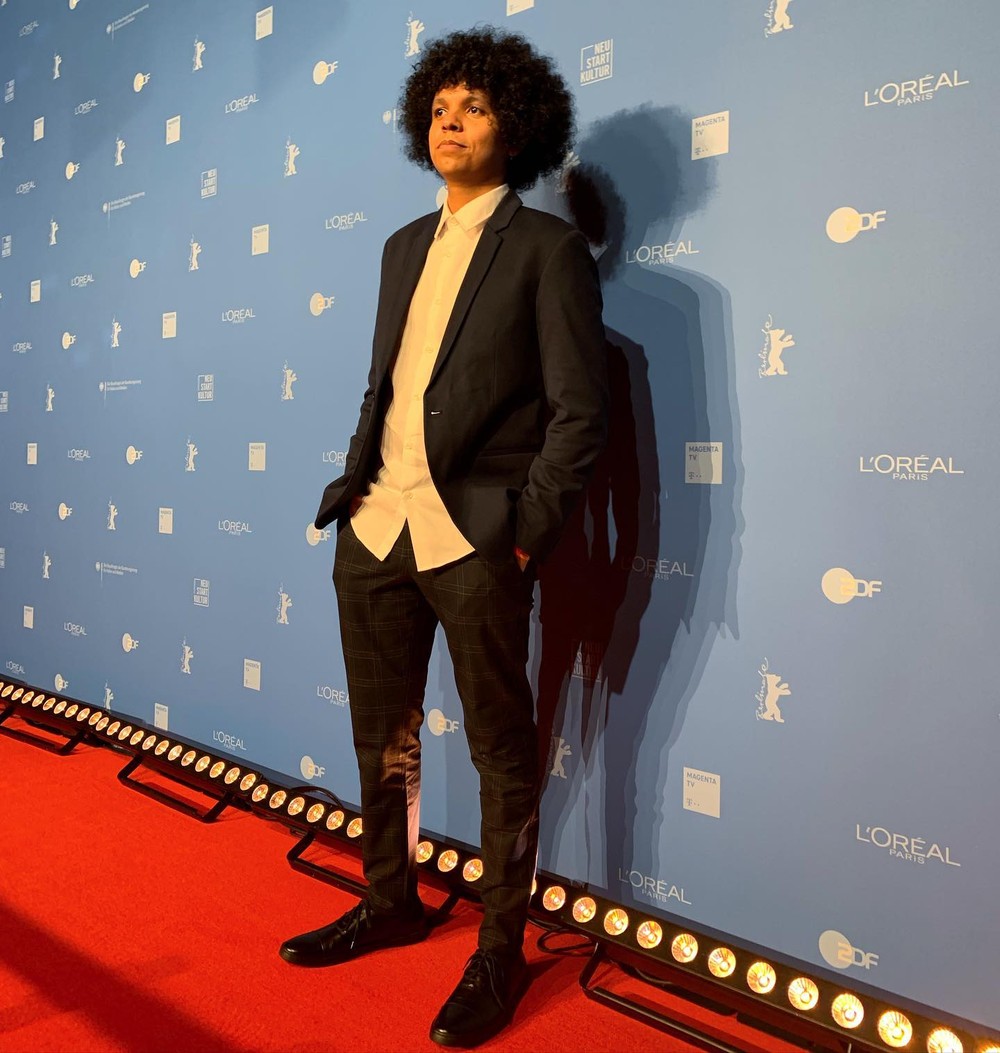 bruno ribeiro O cineasta Bruno Ribeiro usa calça cinza xadrez, camisa branca, paletó cinza e sapato preto. Ele tem cabelo black power e está com as mãos nos bolsos. Está sobre o tapete vermelho e ao fundo há uma parede azul-claro.