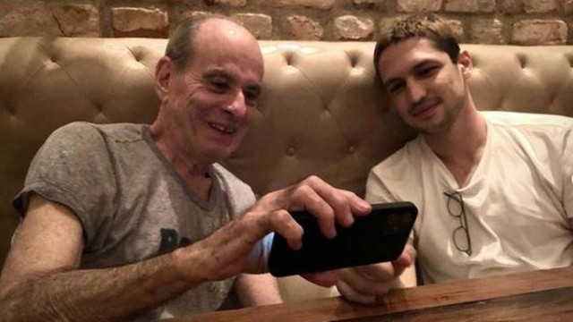 Ney Matogrosso e Gabriel Leone sentados em um sof&aacute; marrom com encosto em capiton&eacute;