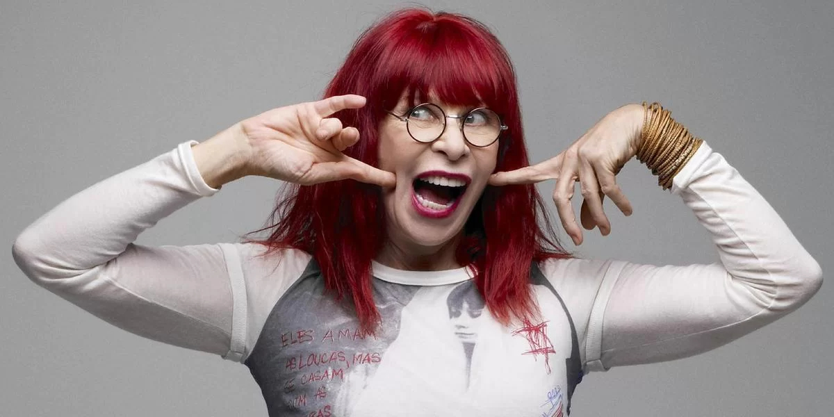 Rita Lee é uma mulher branca, de cabelos ruivos intensos, olhos claros. Ela usa camisa branca e coleto cinza. Está apontando a boca com os dedos.