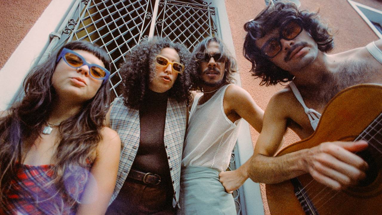 Os cantores Julia Mestre, Dora Morelenbaum, Lucas Nunes e Zé Ibarra, do grupo Bala Desejo, na frente de uma porta de ferro e uma parede rosa. Estão todos de pé e de óculos escuros.