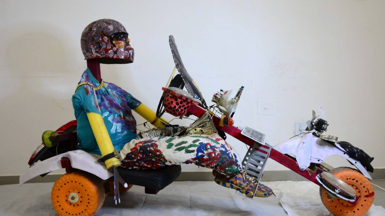Escultura traz figura humana de capacete sentada em um carrinho que lembra um kart.