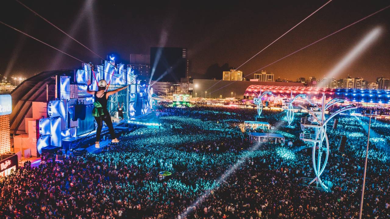 Foto da multidão diante do Palco Mundo do Rock in Rio, com luzes azuis a acesas e vários spots. No canto esquerdo da foto, uma pessoa anda de tirolesa.