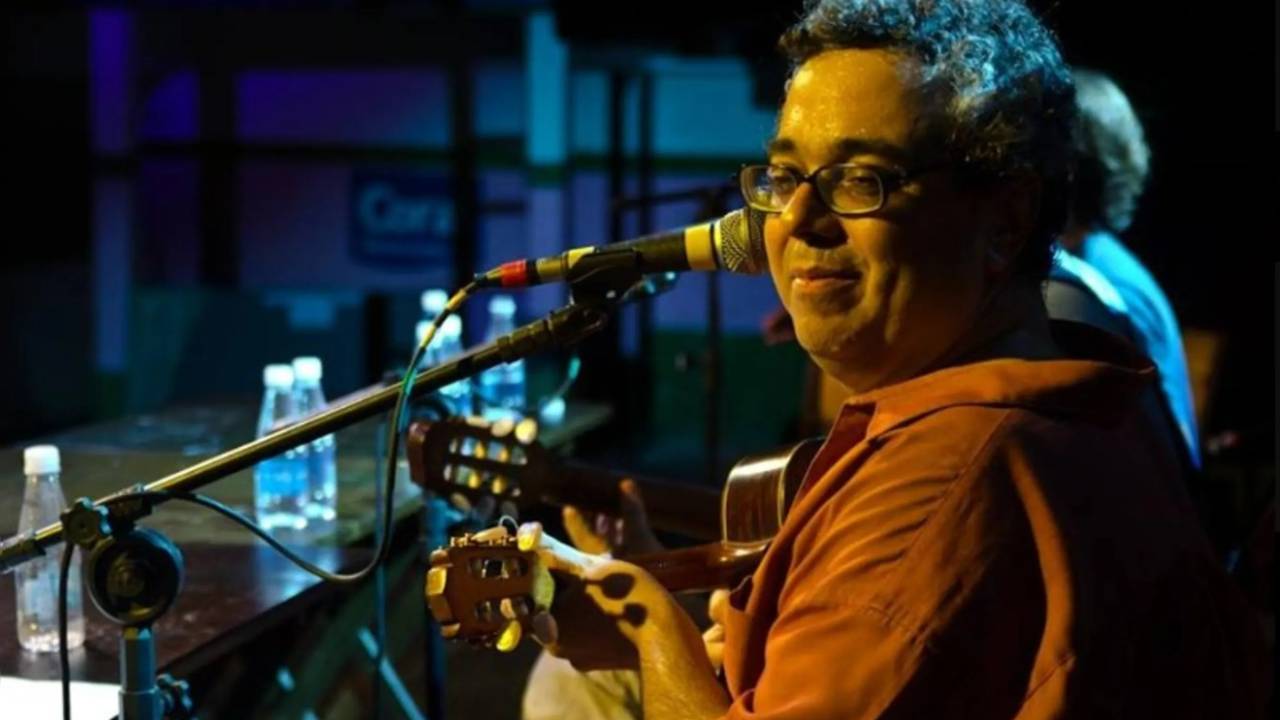 Eduardo Gallotti é branco e tem cabelos cacheados grisalhos. Ele usa óculos, está sentado ao violão (ao fundo, nota-se que é um show, com outros músicos), usa camisa laranja. Está com um leve sorisso.