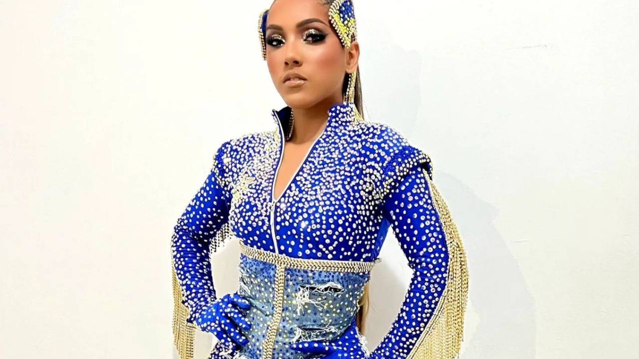 Foto mostra Mayara Lima posando com fantasia brilhosa de manga comprida azul e dourada, com franjas douradas no braço. Mayara olha para a frente e faz pose com as mãos na cintura