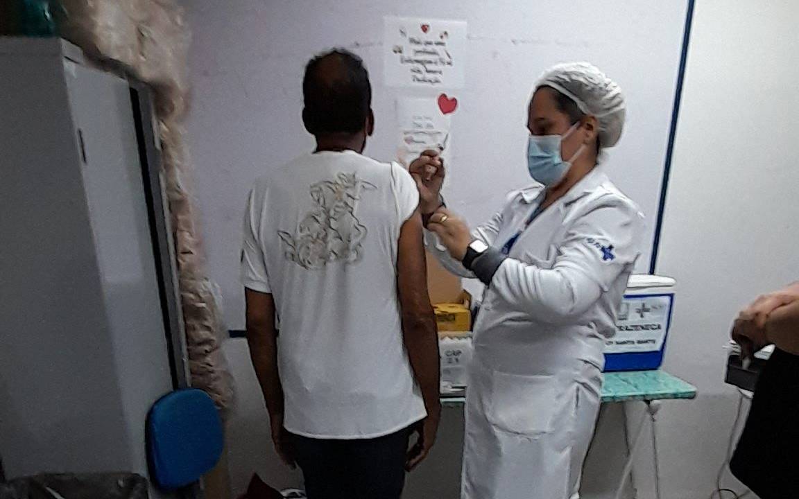 O cantor e compositor Jorge Ben Jor está de costas, vestindo calça jeans e blusa branca com estampa de São Jorge, enquanto é vacinado pela enfermeira do posto de saúde, que está de lado, vestida de branco, usando touca e máscara.