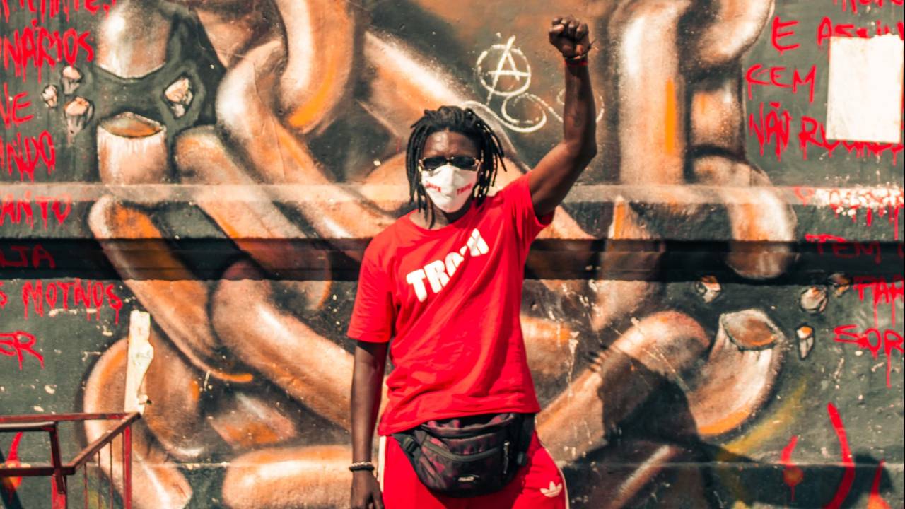 Schackal é um homem negro de tranças curtas. Está de calça Adidas e camiseta com a palavra Tropa escrita, ambas vermelhas. Está de máscara branca e com o punho erguido. Ao fundo, um grafite com correntes.