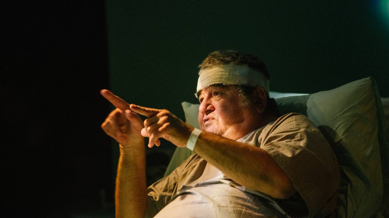 O ator Otávio Augusto está apoiado em uma cama, gesticulando, e tem uma atadura em torno da testa.