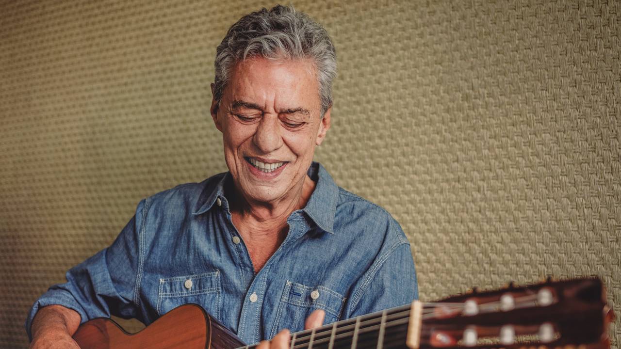 Chico Buarque está de camisa jeans com as mangas dobradas, segurando o violão. Ele olha para baixo e sorri.
