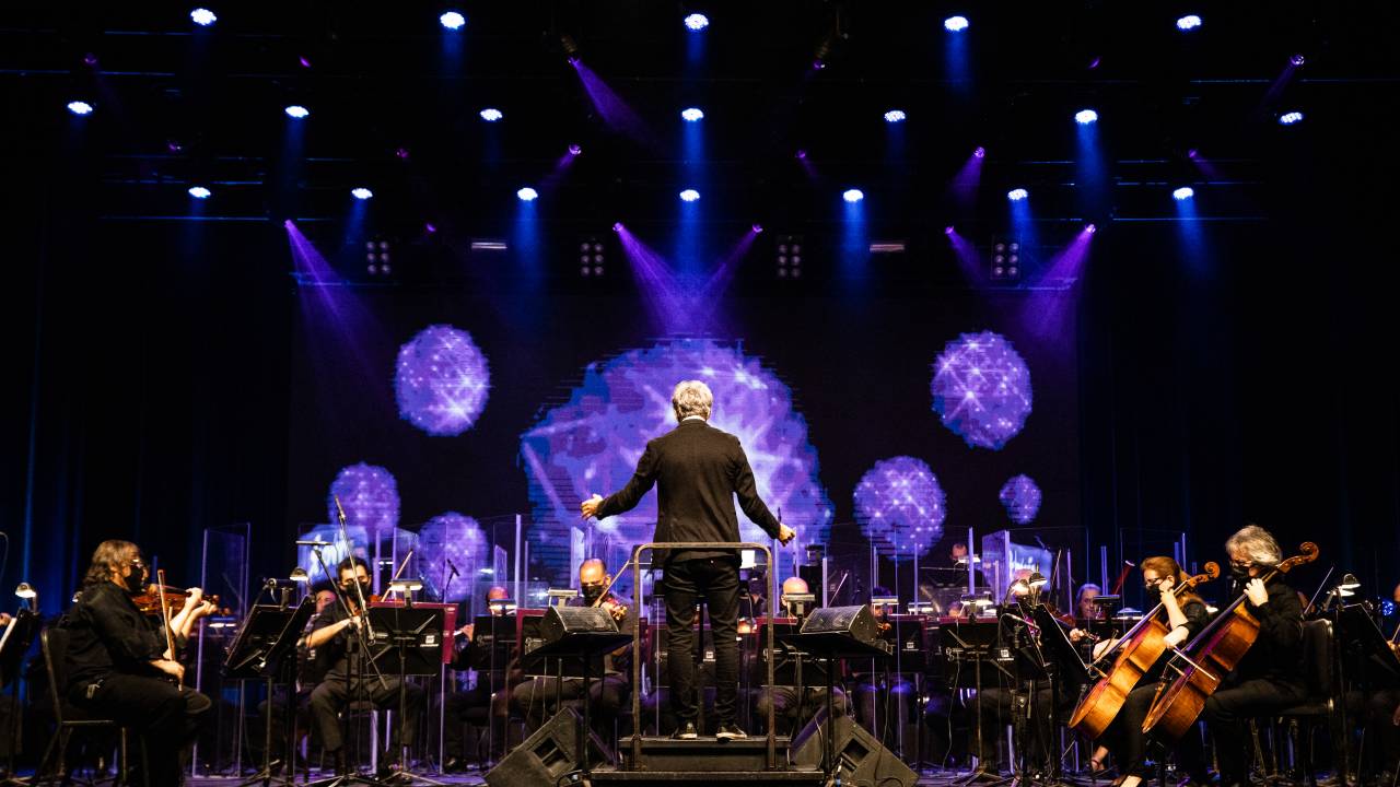 Fotos do maestro de costas regendo a orquestra, com projeções roxas na parede.