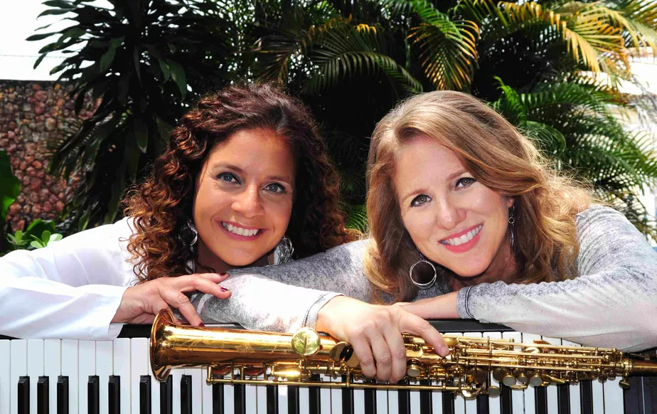 À esquerda, a pianista Sheila Zagury, branca de cabelos castanhos cacheados, apoiada em um teclado. À direita, Daniela Spielmann, branca e loura, segurando uma flauta transversal. As duas estão sorrindo.