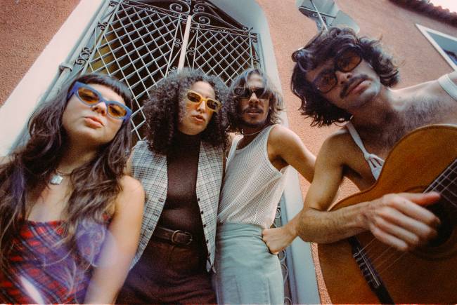 A partir da esquerda, o coletivo Bala Desejo: Julia Mestre, Dora Morelenbaum, Lucas Nunes e Zé Ibarra, fotografados de baixo para cima, em frente ao portão de um casarão de parede rosa-claro.